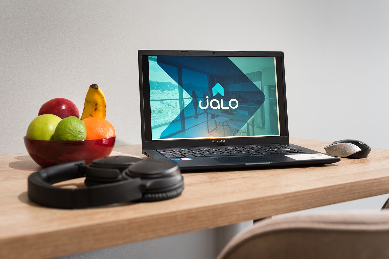 A laptop on a wooden desk with a bowl of fruit, a pair of headphones, and a mouse.