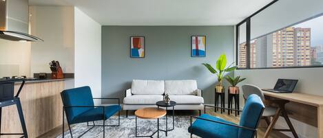 Una sala de estar moderna con un sofá, dos sillones azules, una mesa de centro y un escritorio con una computadora portátil. Hay pinturas abstractas en la pared y un ventanal con vista a la ciudad.