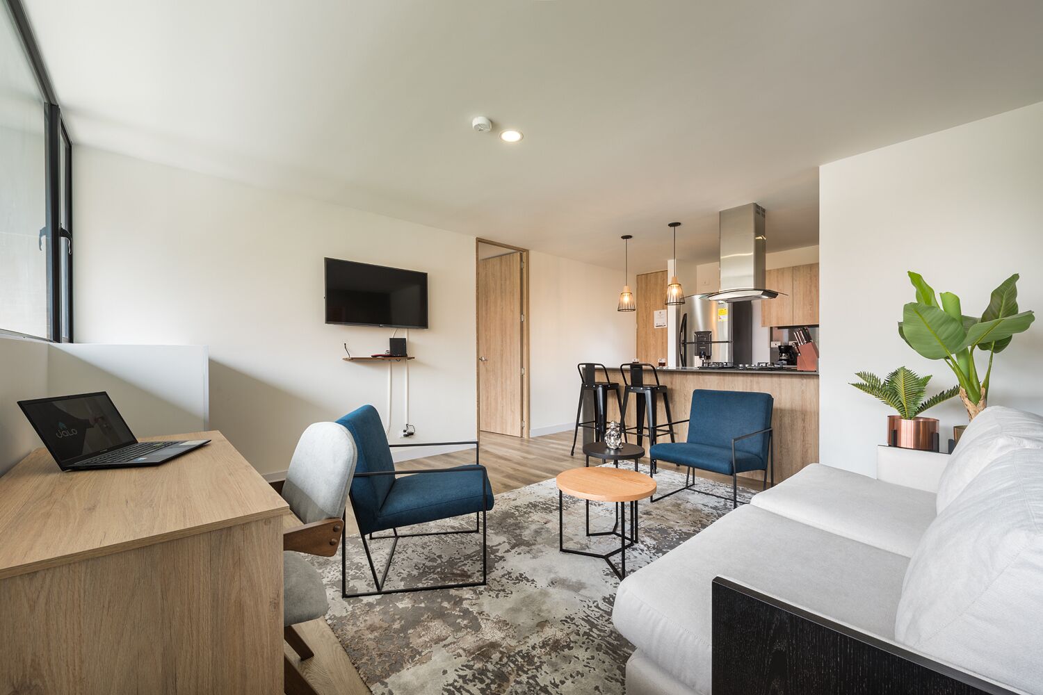 A modern apartment with a living room, dining area, and kitchen. There is a flat-screen TV mounted on the wall, a laptop on a desk, and a sofa.