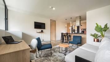 A modern apartment with a living room, dining area, and kitchen. There is a flat-screen TV mounted on the wall, a laptop on a desk, and a sofa.