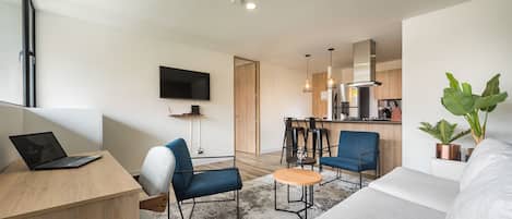 Un apartamento moderno con sala de estar, comedor y cocina. Hay un televisor de pantalla plana montado en la pared, una computadora portátil sobre un escritorio y un sofá.