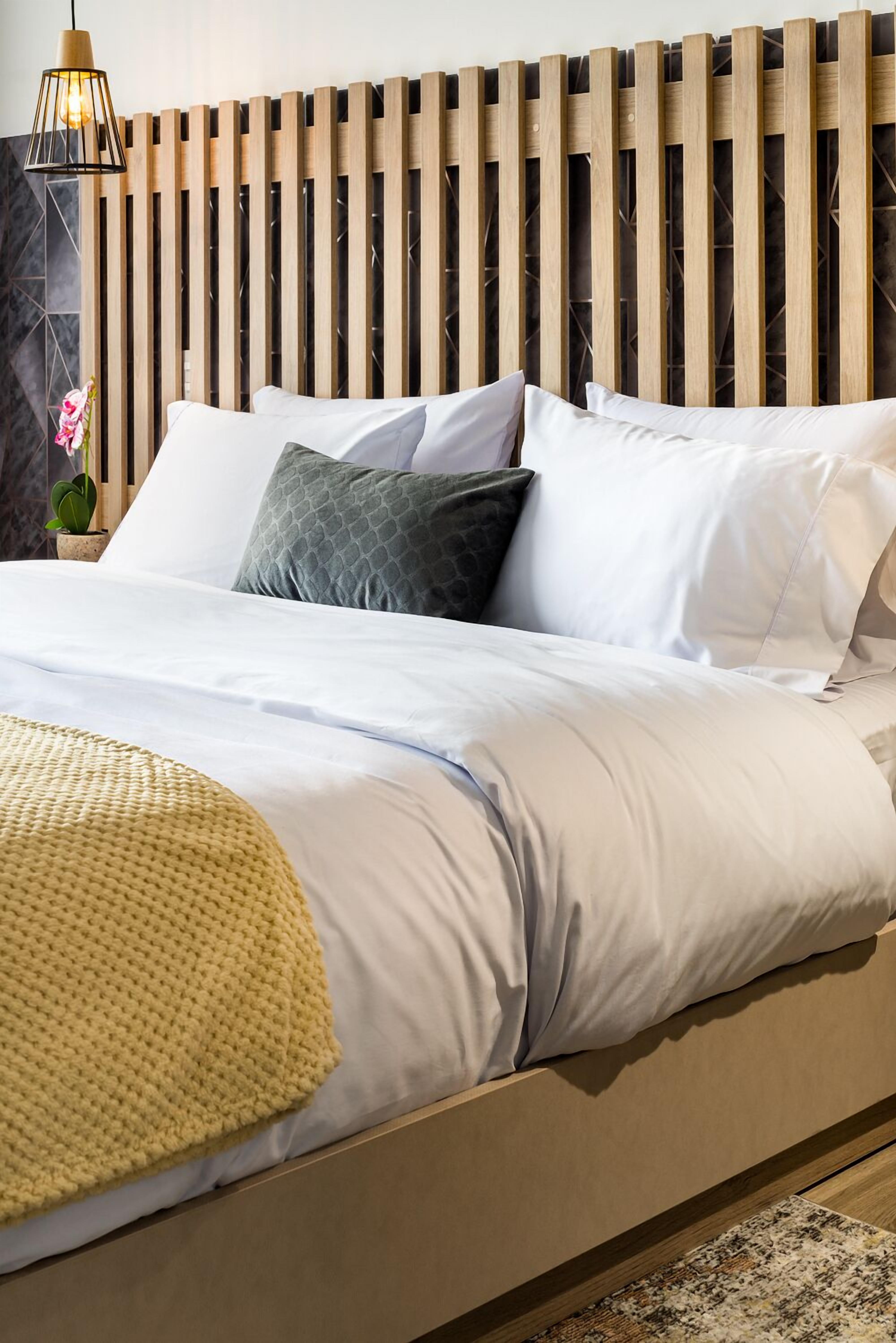 Een bed met wit en grijs beddengoed, een gele deken en een houten hoofdbord.