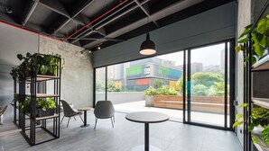 A modern office space with a round table, two chairs, a bookshelf with plants, and large windows offering a view of buildings and greenery. - Go Living and Suites by Jalo (Medellín)