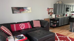 Een woonkamer met een zwarte leren bank, rode en witte kussens, een rood-wit kleed en een keuken op de achtergrond.