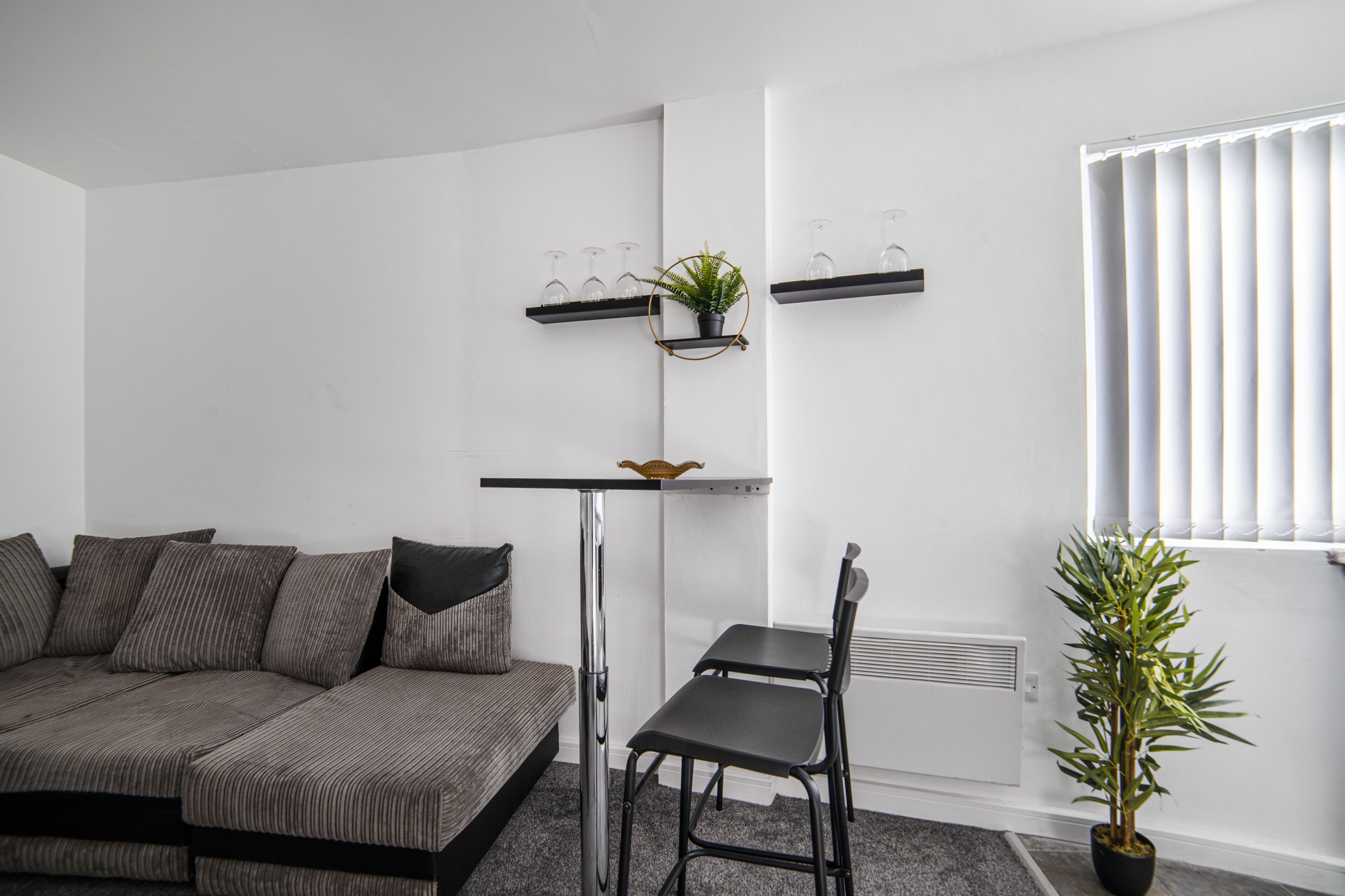 A modern living room with a sectional sofa, a small table, and two chairs. There are two shelves with decorative items and a potted plant.