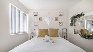 Een net opgemaakt bed met witte lakens en twee gouden kussens, een nachtkastje met een lamp, en een grote spiegel aan de muur.