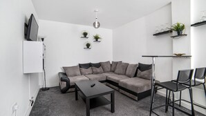 Een moderne woonkamer met een modulaire bank, een salontafel en twee barkrukken. Er staan drie potten met planten op de muur, een flatscreen televisie is aan de wand bevestigd, en er hangt een hanglamp.