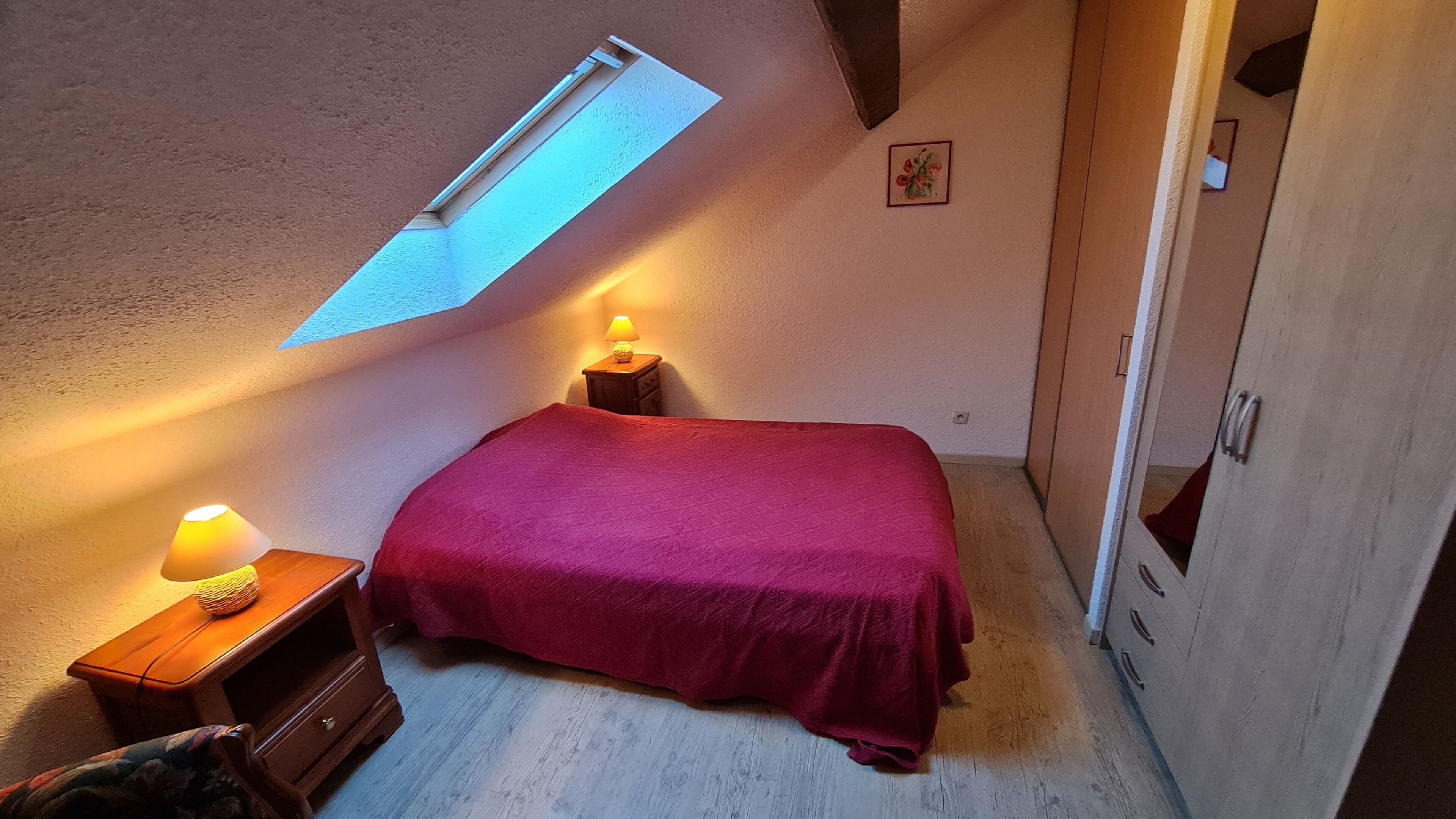 Une chambre avec un puits de lumière, un lit recouvert d’une couverture rouge, une table de chevet avec une lampe et une armoire.