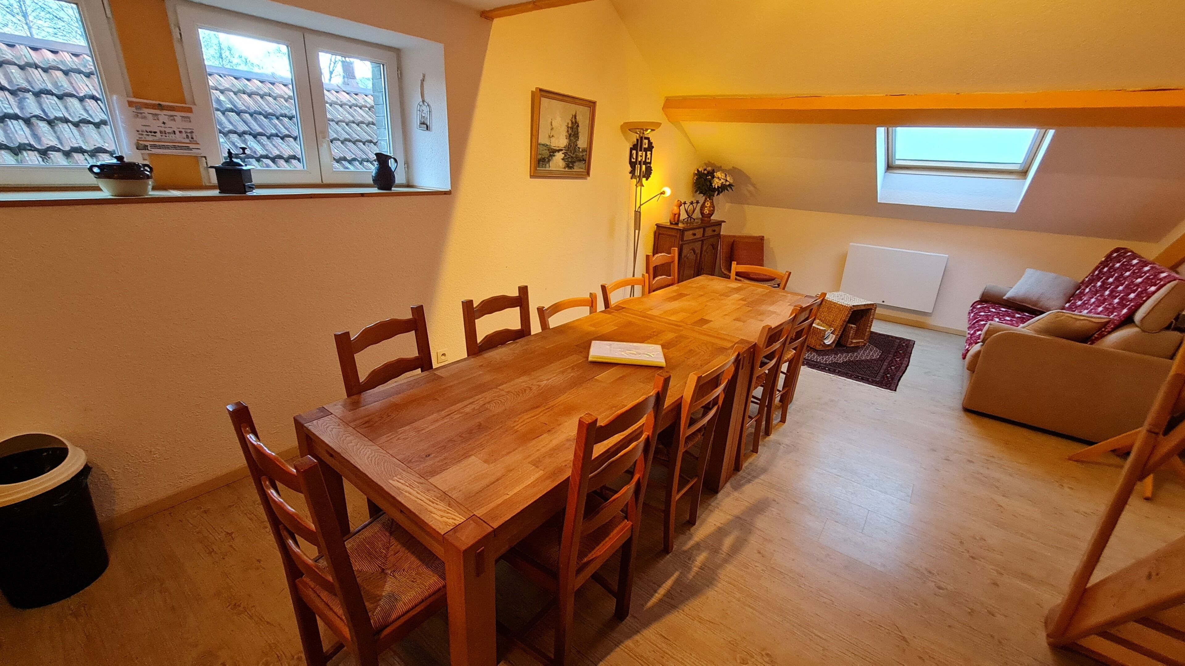 Un comedor con una mesa y sillas de madera, un sofá, una ventana con vistas a un tejado de tejas y una lámpara.