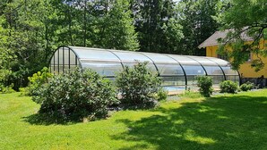 A greenhouse structure with a transparent roof, surrounded by lush greenery and a yellow building in the background. - DOMAINE DES GRAVIERS (Plainfaing)
