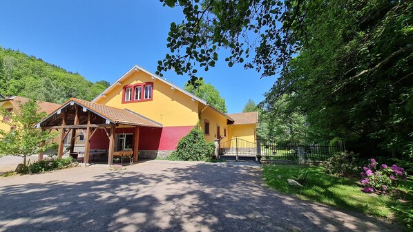 Un bâtiment jaune avec un toit rouge, entouré de verdure et bordé d’une allée en gravier.