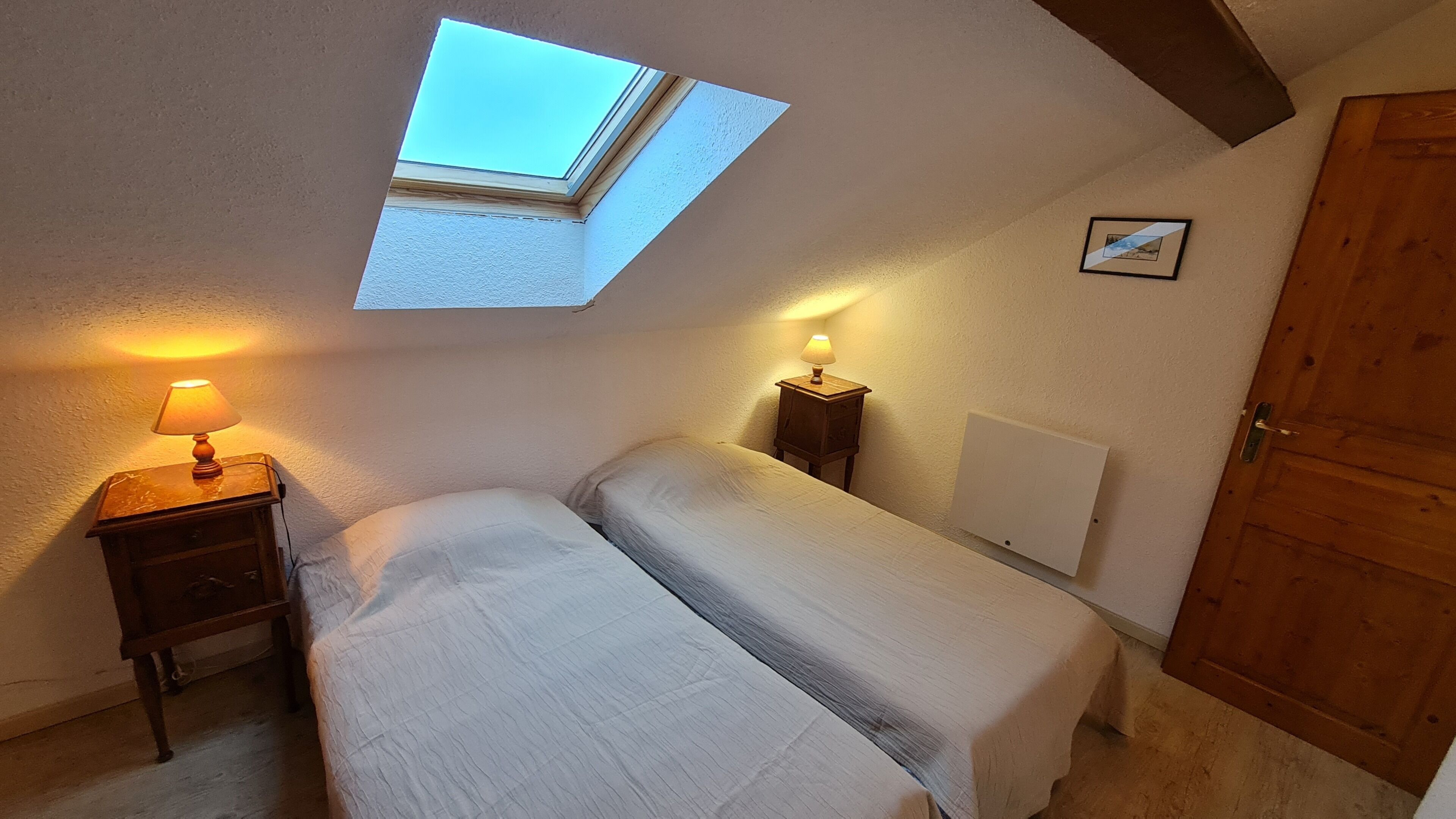 A bedroom with a skylight, a bed, bedside tables with lamps, a wooden wardrobe, and a framed picture on the wall.