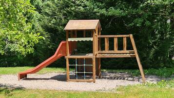 Une structure de jeu en bois, dotée d’un toboggan et d’une zone d’escalade, entourée de gravier et de verdure.