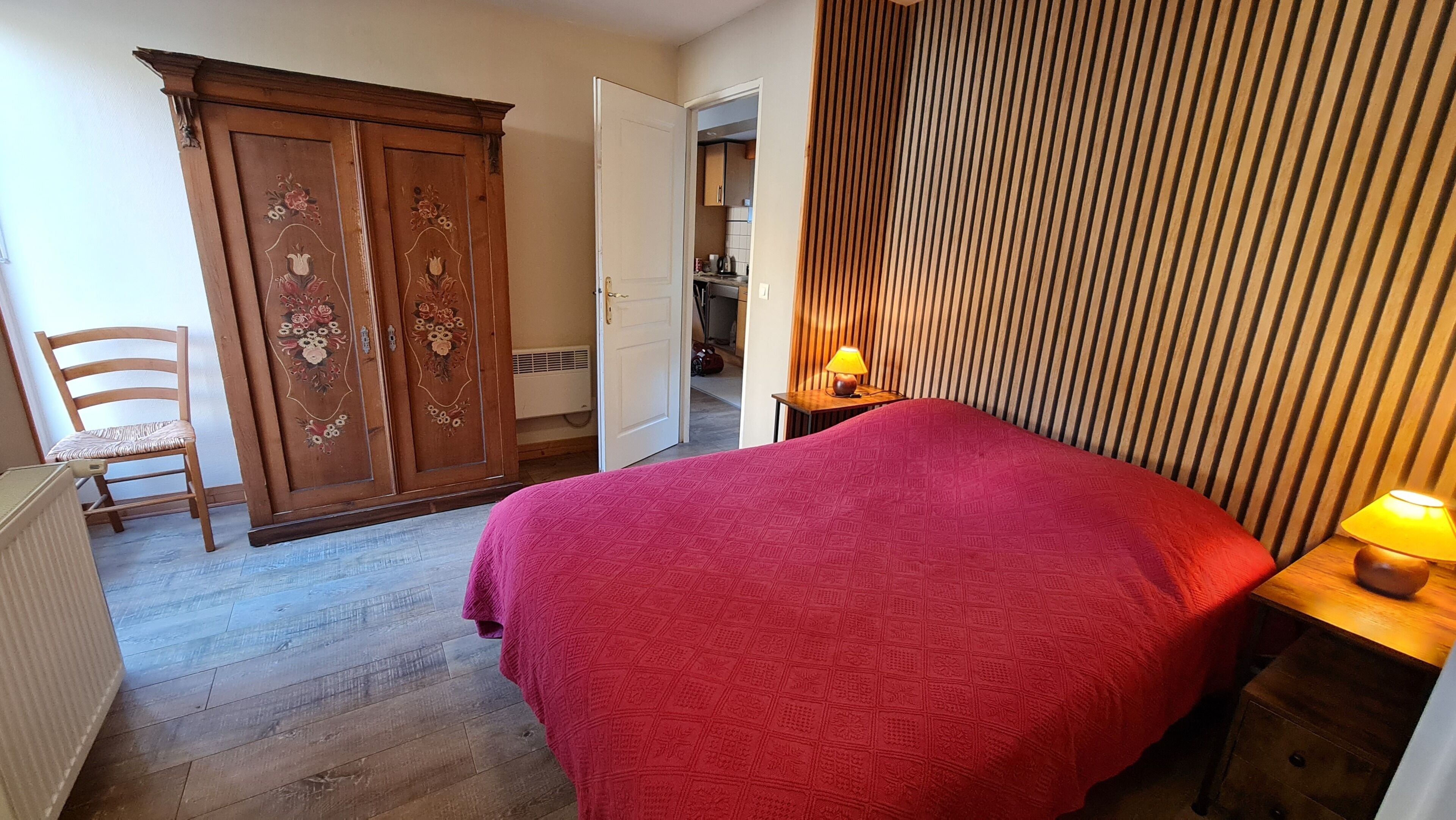 A bedroom with a bed, a wooden wardrobe, a chair, and a bedside table with a lamp.