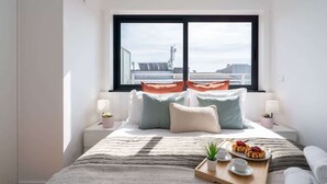 A neatly arranged bedroom with a large window, a bed with multiple pillows, bedside tables with lamps, and a tray with food and drinks. - Bright Flat w/ Balcony |  Ace Location by HostWise (Porto)