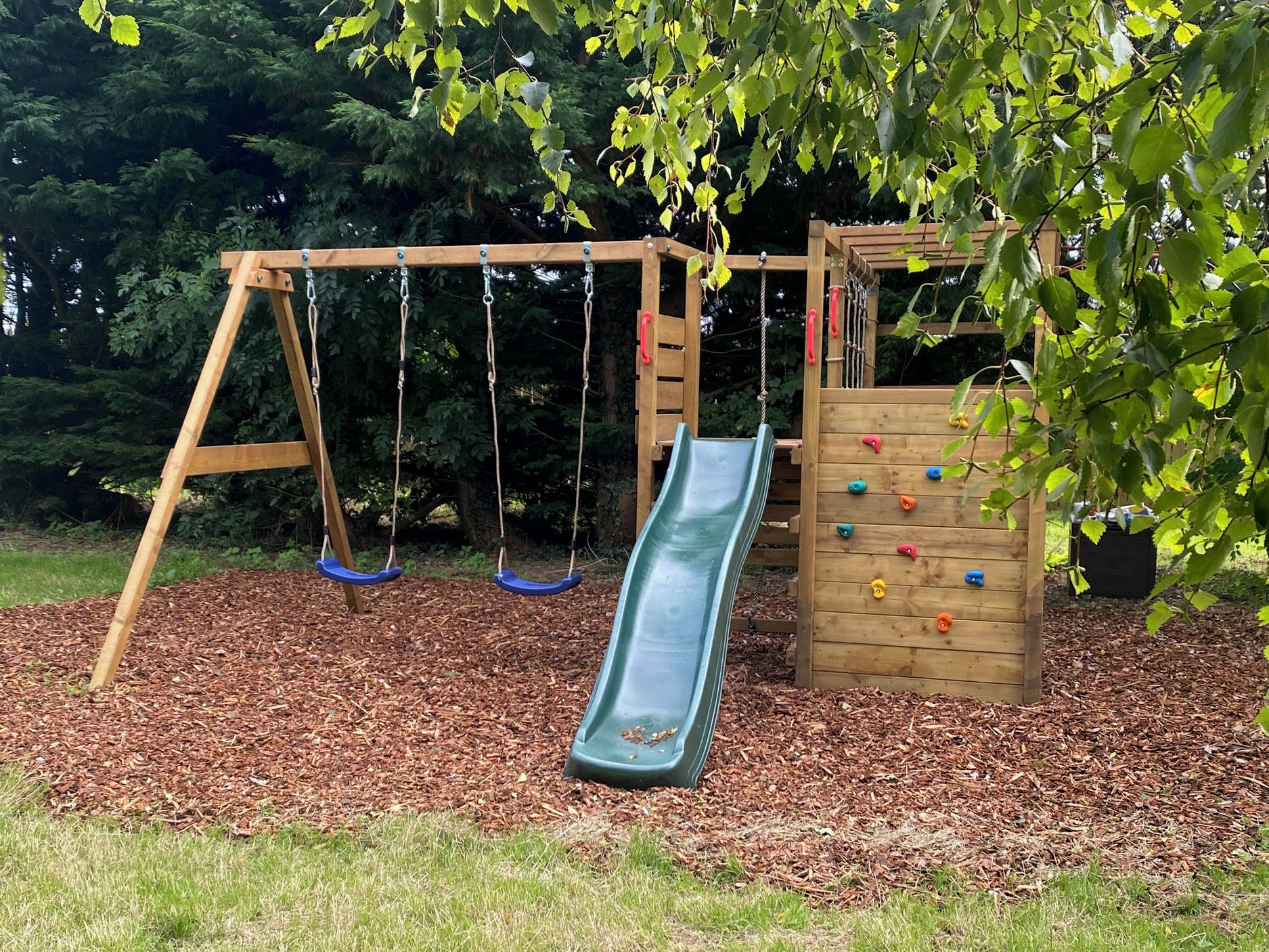 A wooden swing set with a slide, surrounded by mulch and trees.