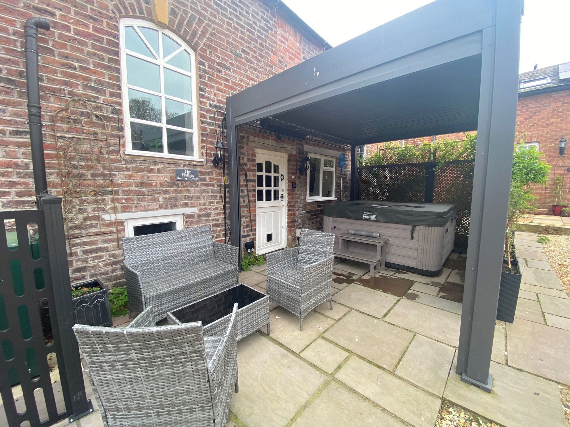 A covered patio with wicker furniture, a brick house, and a sign that reads 'The Hollies Holiday Cottage.'
