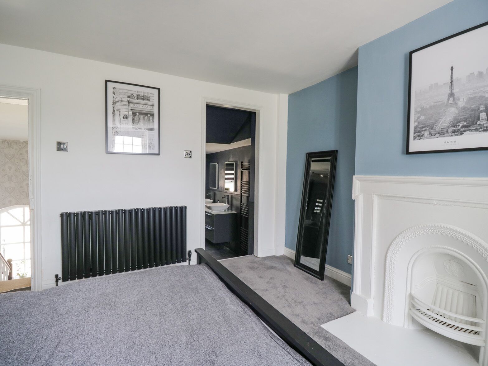 Een slaapkamer met een bed, een radiator, een spiegel, een open haard en een deur naar een badkamer.