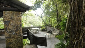 Une terrasse en bois, soutenue par des piliers en pierre, entourée d’une végétation luxuriante et d’arbres.