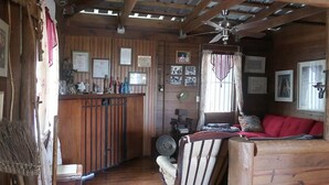 L’intérieur d’un chalet rustique, avec un plafond en bois, un lit recouvert d’une couverture rouge, un bureau en bois sur lequel se trouvent divers objets, et un ventilateur.