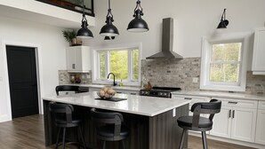Una cucina moderna con isola centrale, sgabelli da bar, illuminazione a sospensione e vista sull'esterno attraverso le finestre.