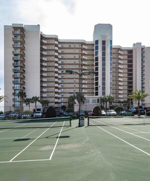 A tennis court with a net in the middle, surrounded by a parking area and palm trees.