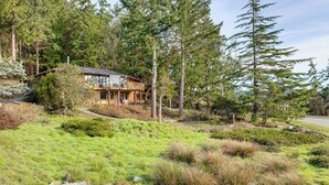 A modern house with a large deck, surrounded by dense forest and grassy terrain. - Serene La Conner Retreat w/ Fireplace & Deck! (La Conner)