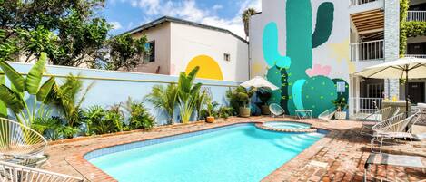 A swimming pool with a brick border, surrounded by lounge chairs and umbrellas, with a mural of cacti and a sun on the adjacent wall.