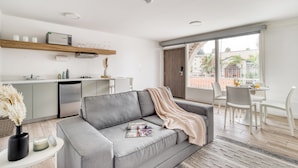 Una sala moderna con un sofá gris, una mesa de centro y un comedor con sillas blancas. Se ve una cocina con microondas y refrigerador al fondo.