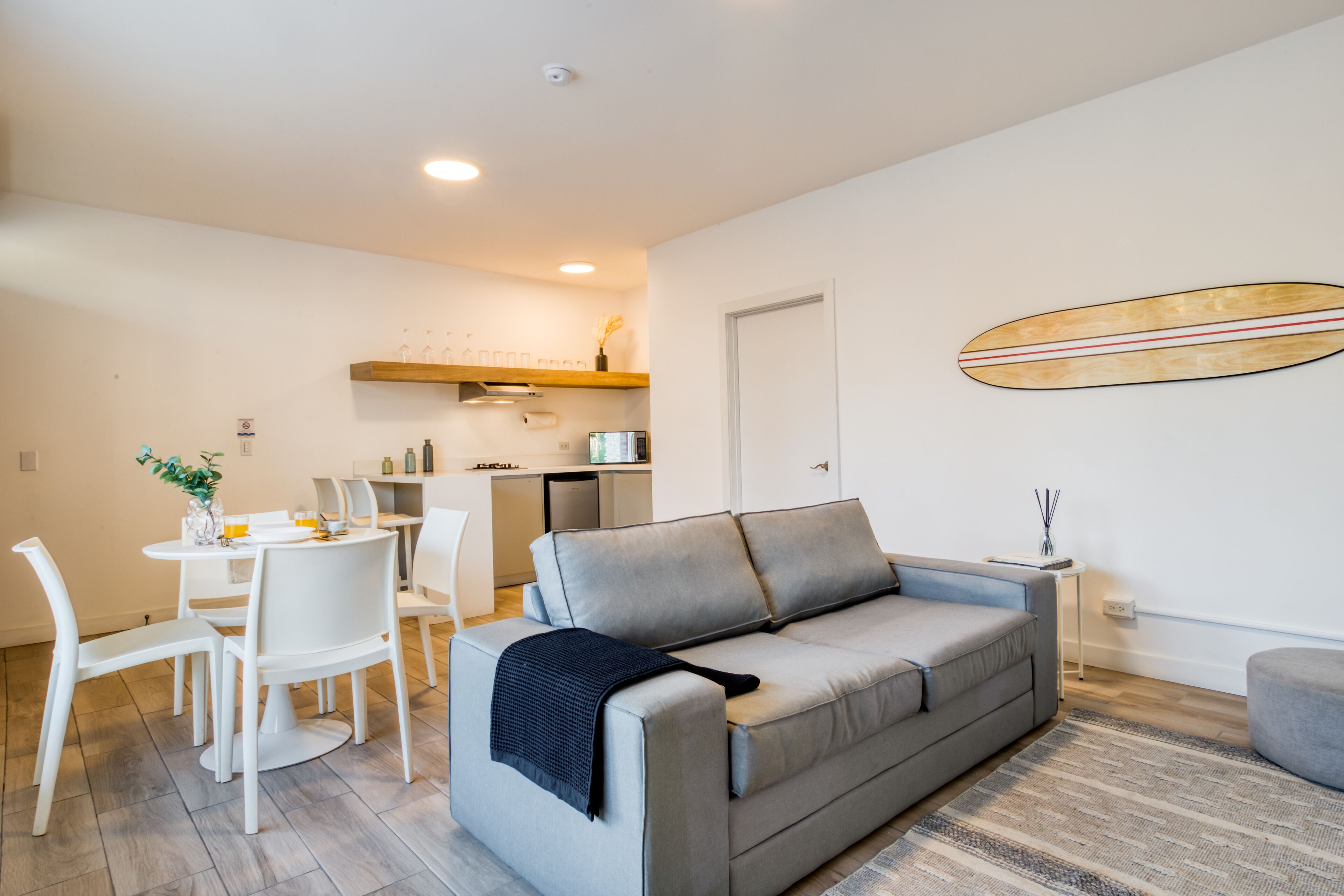 A modern living room with a grey sofa, a dining table with chairs, a kitchen area, and a surfboard decoration on the wall.