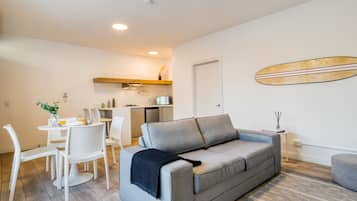 A modern living room with a grey sofa, a dining table with chairs, a kitchen area, and a surfboard decoration on the wall.