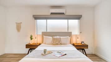 A modern bedroom with a large bed, bedside tables, a wall-mounted air conditioner, and a window with blinds.