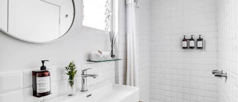 Un baño con lavamanos blanco, espejo y ducha. Hay artículos de aseo en la pared y una planta en el mostrador.
