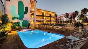Una piscina con sillas de descanso y sombrillas, un mural de cactus y un edificio de varios pisos con balcones.