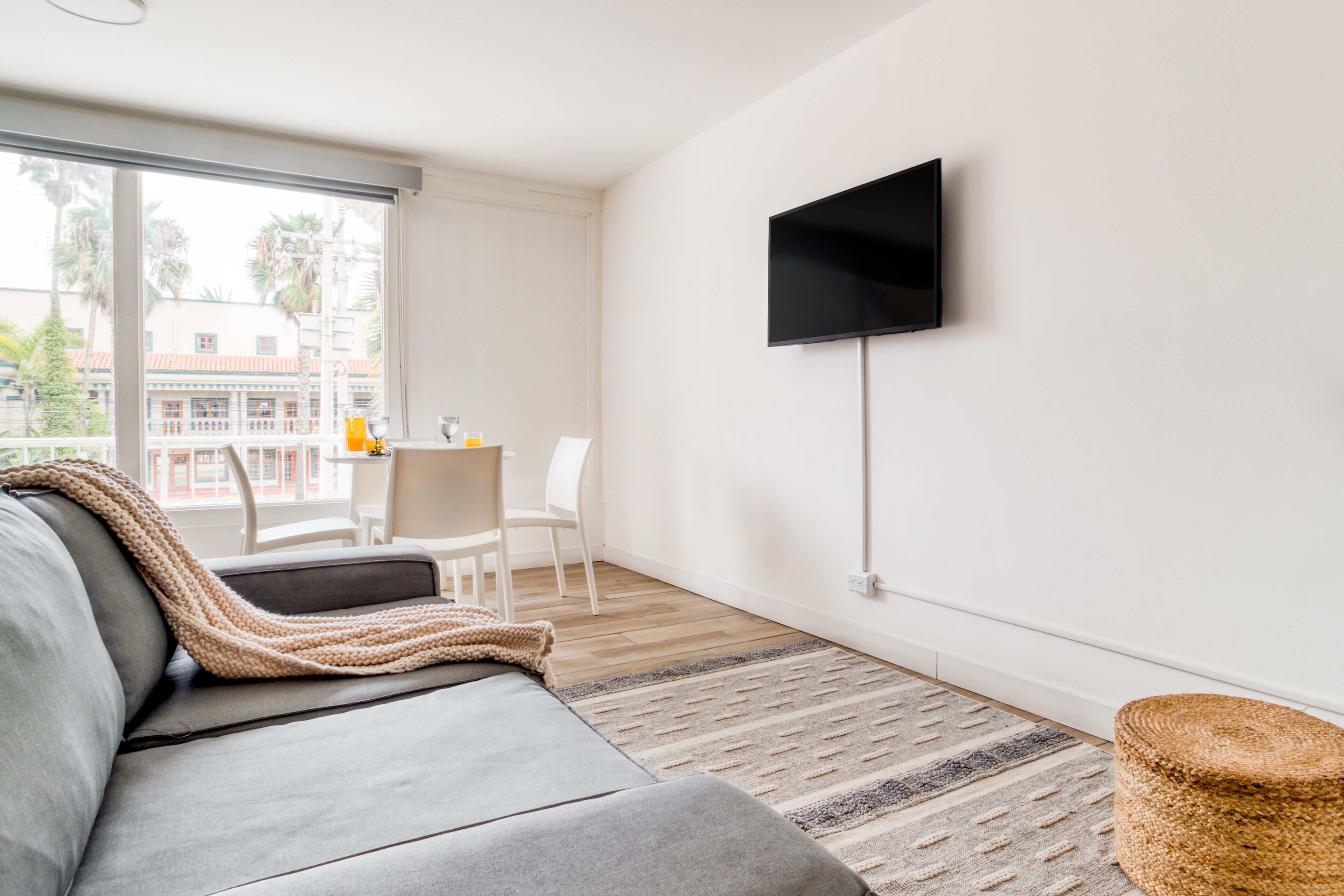 A modern living room with a grey sofa, a flat-screen TV mounted on the wall, a small round table with chairs, and a large window offering a view of palm trees and buildings.