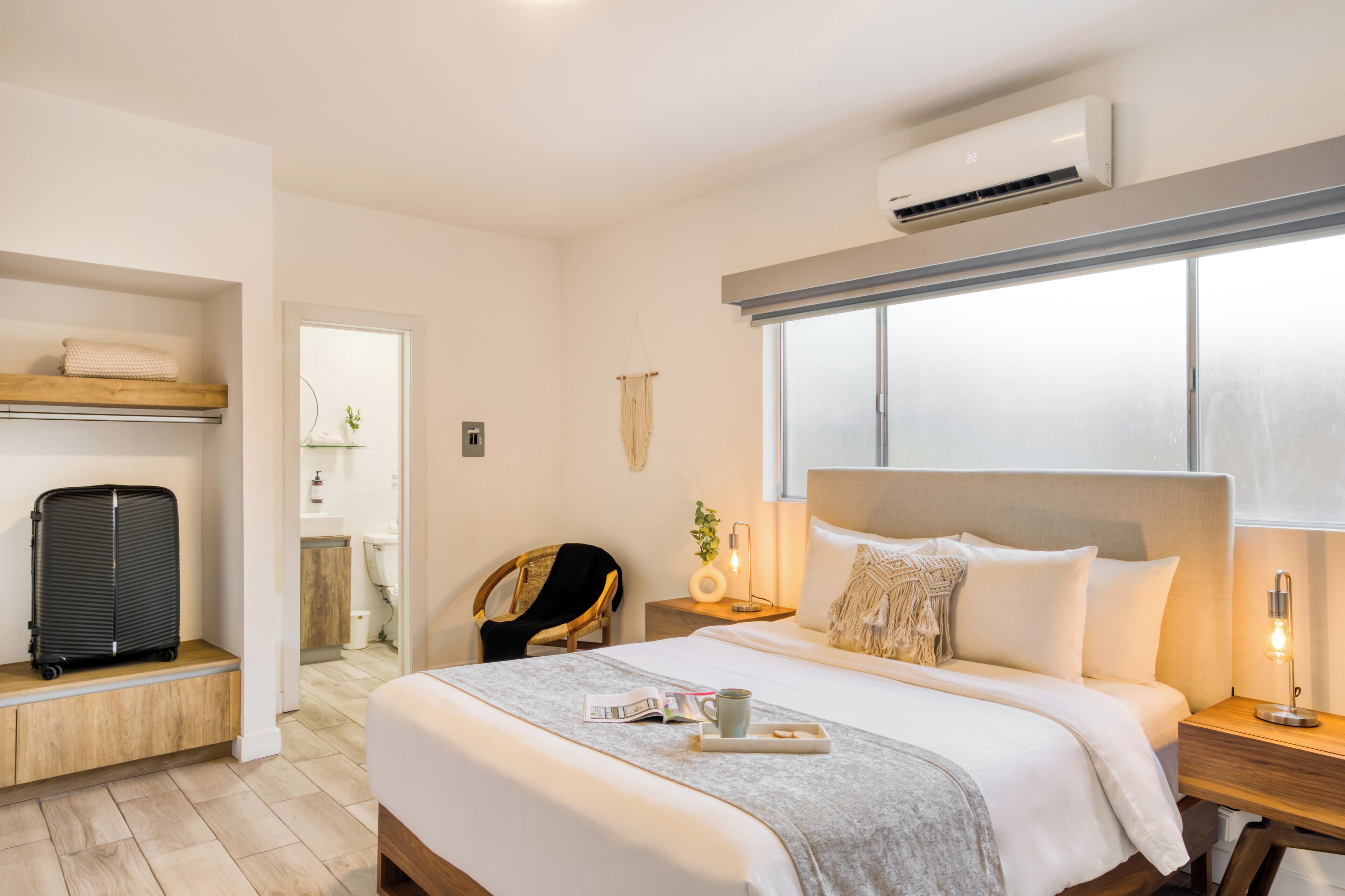 Une chambre d’hôtel moderne avec un grand lit, une table de chevet avec une lampe, une valise posée sur une étagère et un appareil de climatisation.