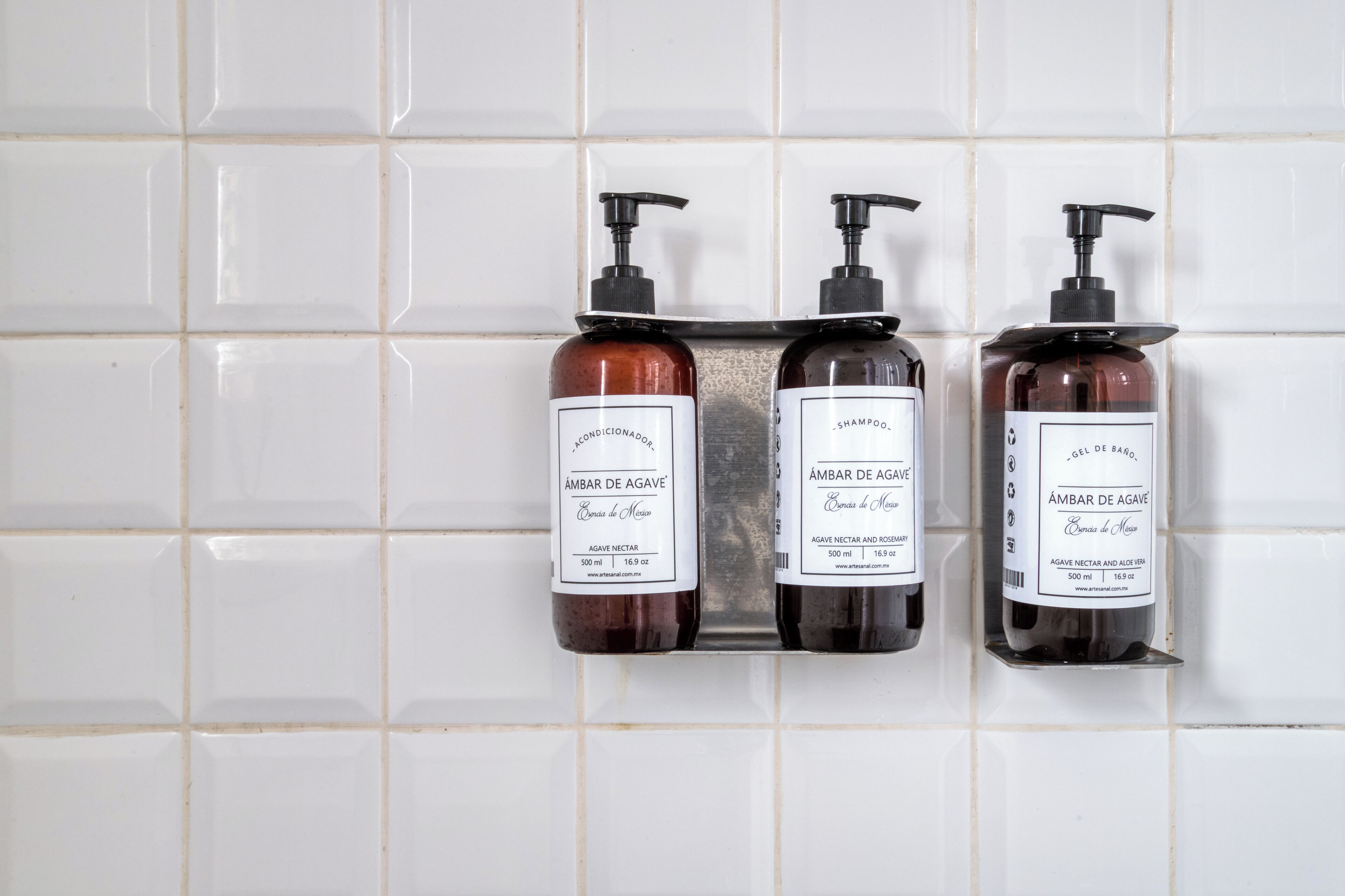 Tres botellas de champú y acondicionador 'Ámbar de Agave' sobre una pared de azulejos.