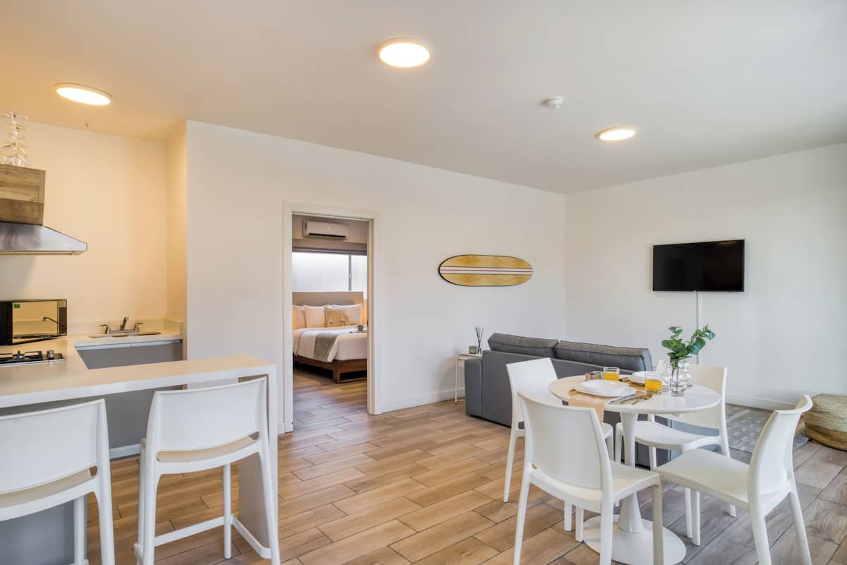 A modern, well-lit room with a kitchen area, a dining table with chairs, a sofa, and a TV mounted on the wall.