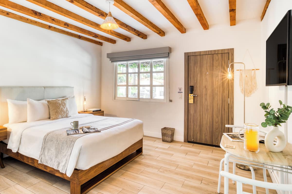 A bedroom with a wooden bed, a bedside table, a window, a door, and a small table with a plant and a glass of orange juice.