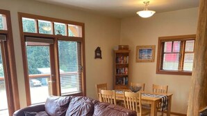 A living room with a brown leather sofa, a patterned armchair, a wooden dining table with chairs, a bookshelf, and large windows with blinds. - Red Rob #6 - 2 bedroom Sleeps 6 (Rossland)