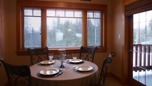 A dining area with a round table set for four, flanked by two windows with blinds, and a door with a metal gate. - Redstone Chalets C Red Mountain (Rossland)
