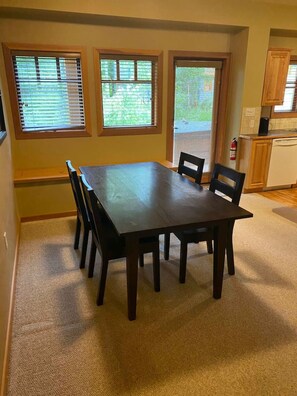 Comedor con mesa y sillas de madera oscura, tres ventanas con persianas, zona de cocina con armarios de madera y un extintor de incendios en la pared.