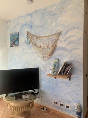 A room with a television on a glass table, a wall with a painted seascape, a shelf with magazines, and a power outlet.