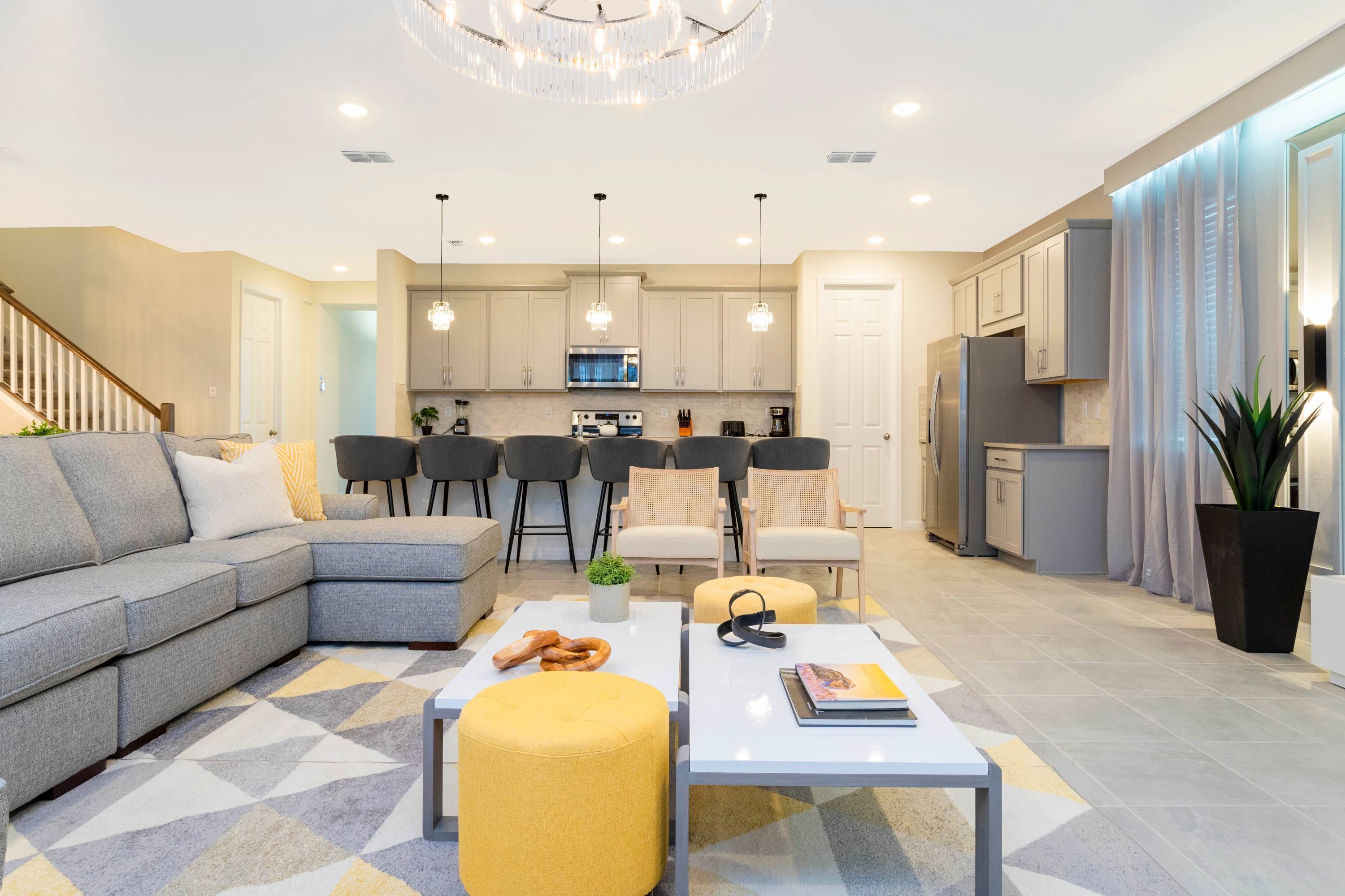 A modern living room with a grey sectional sofa, a yellow ottoman, and a white coffee table. There is a kitchen area with white cabinets and a dining area with black chairs.