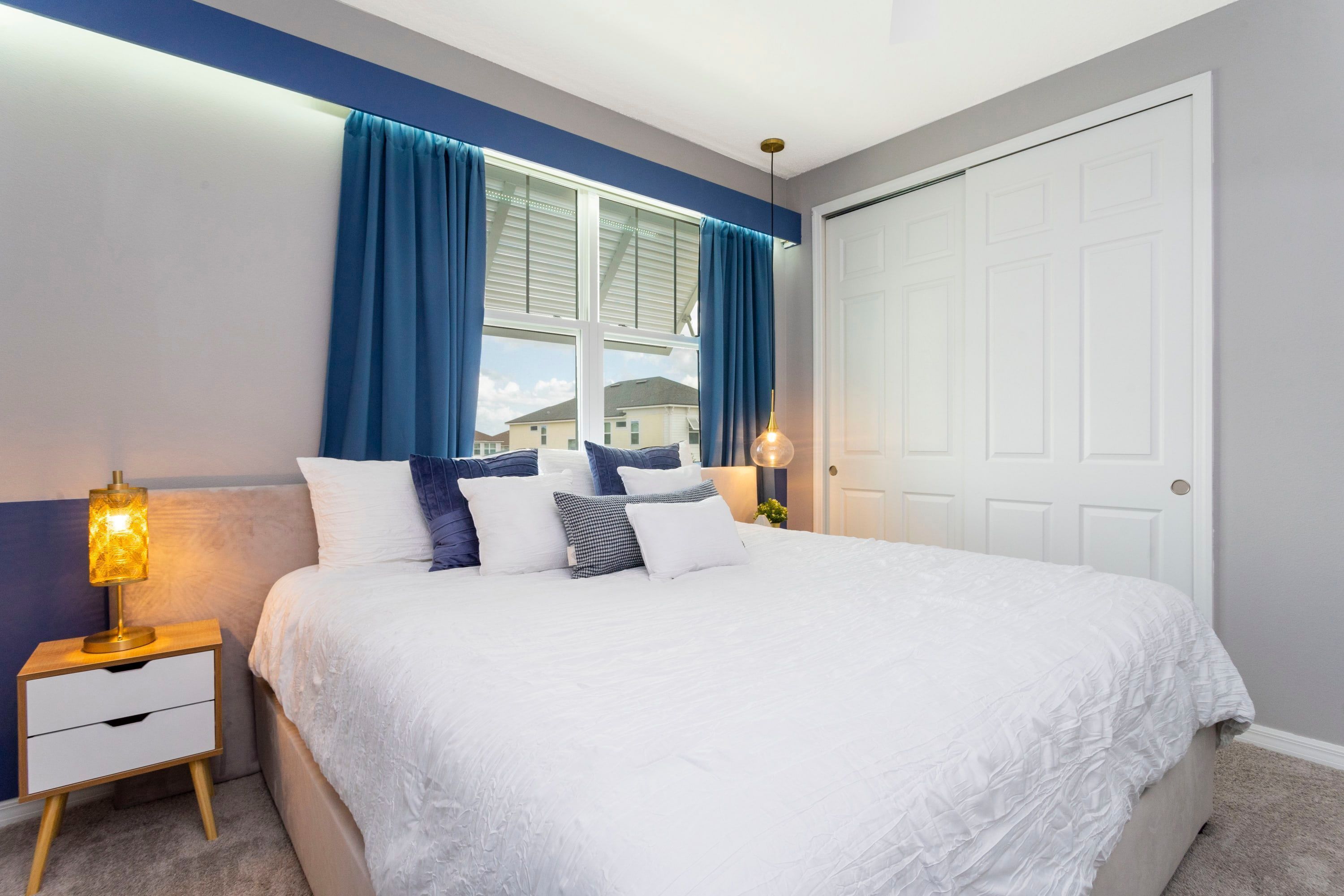 Una cama bien hecha con ropa de cama blanca y varios almohadones, una mesita de noche con una lámpara, y una ventana con cortinas azules.