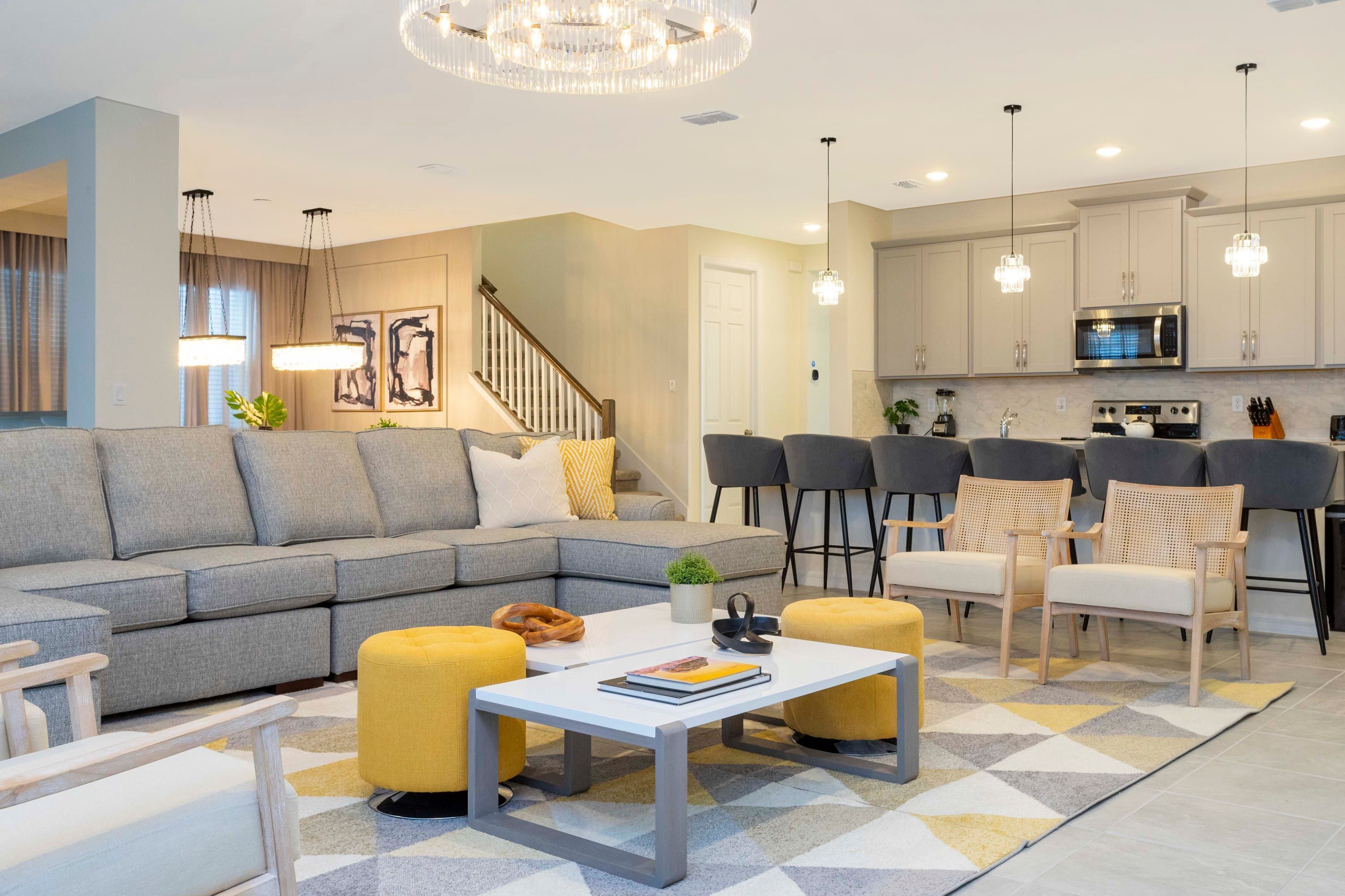 A modern living room with a grey sofa, yellow ottomans, and a white coffee table. There is a staircase leading to another level, and a kitchen area with white cabinets and bar stools.
