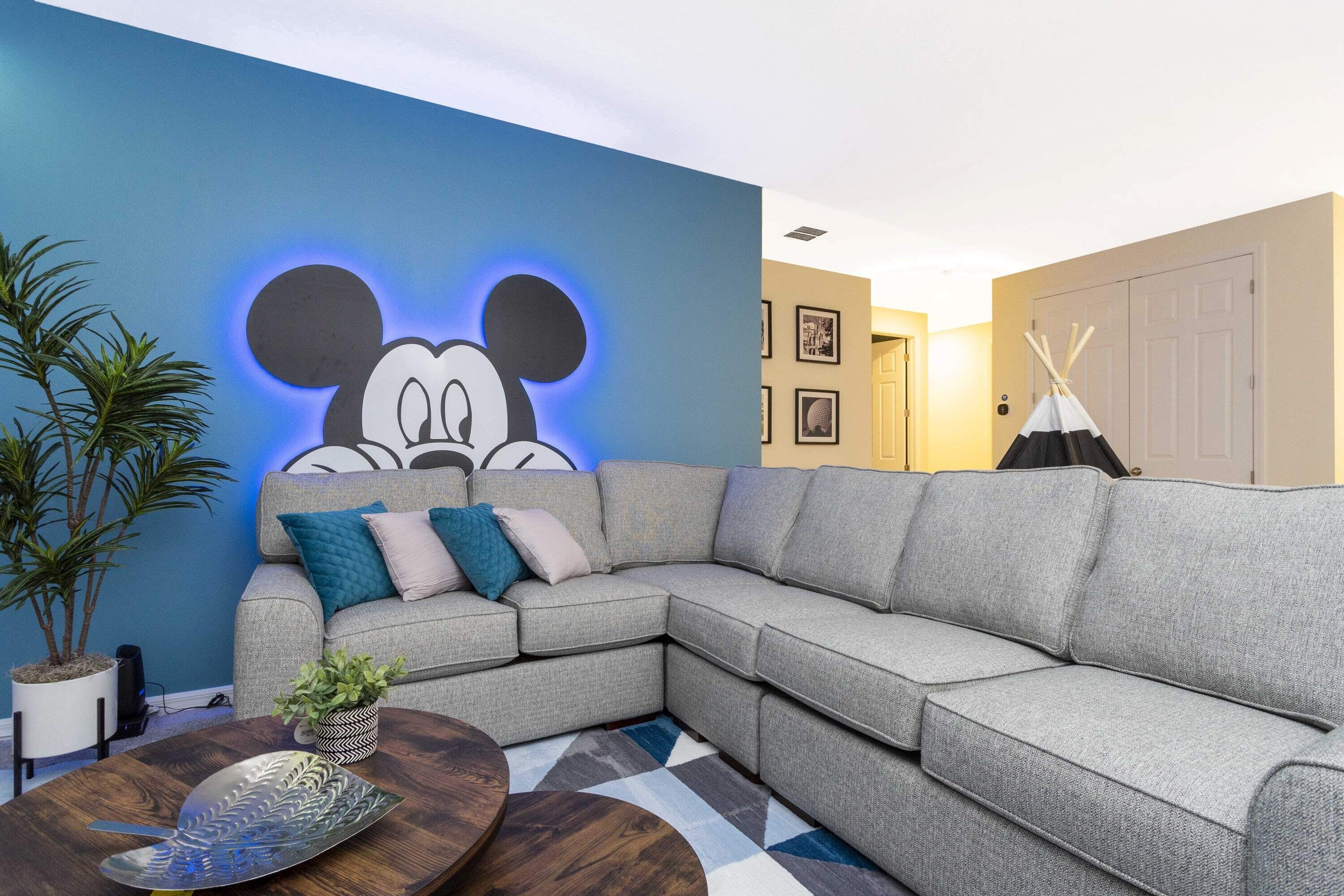 A modern living room with a large sectional sofa, a decorative wall with a Mickey Mouse mural, a potted plant, and a round coffee table.