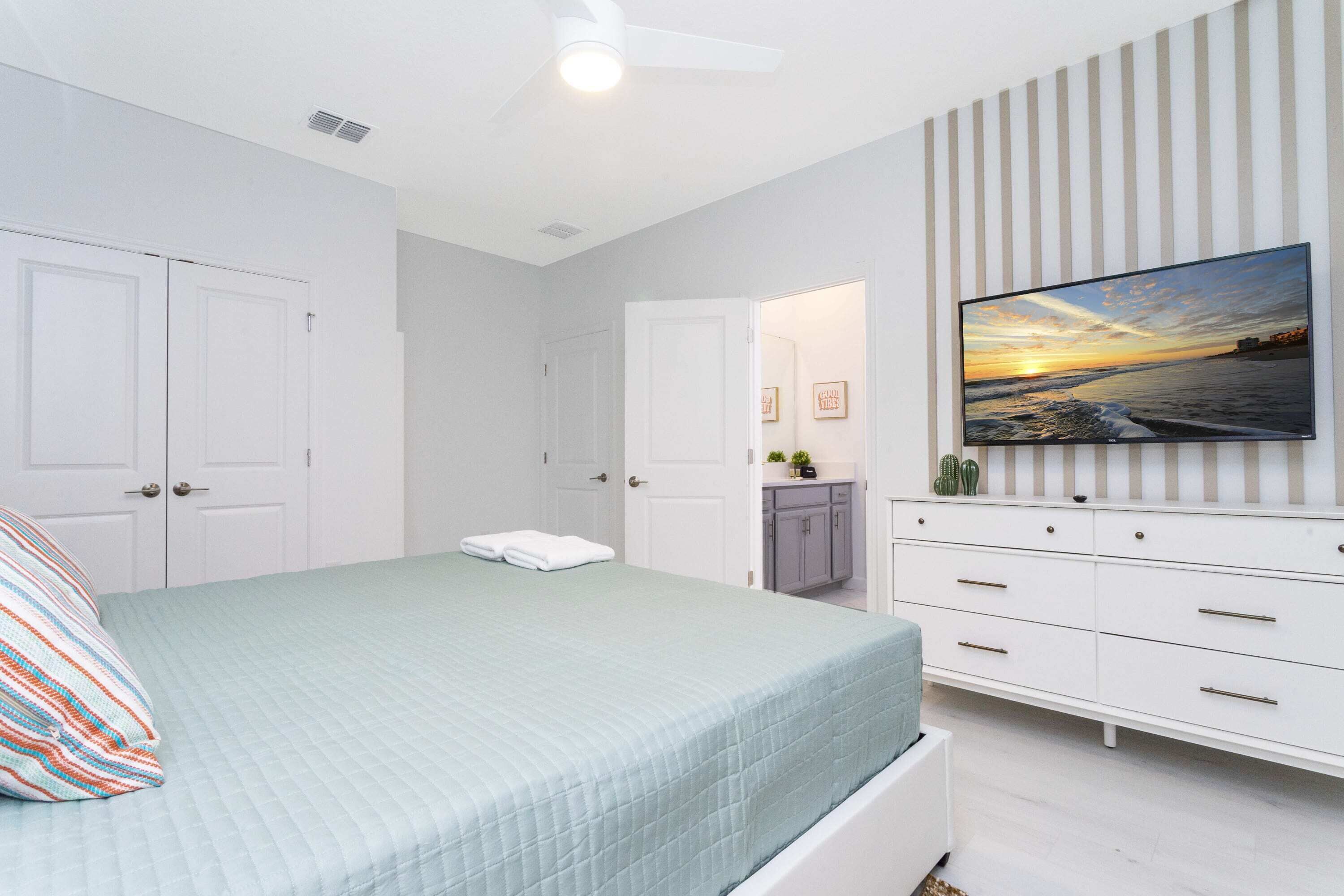 A bedroom with a bed, a TV mounted on the wall, a ceiling fan, and a door leading to another room.