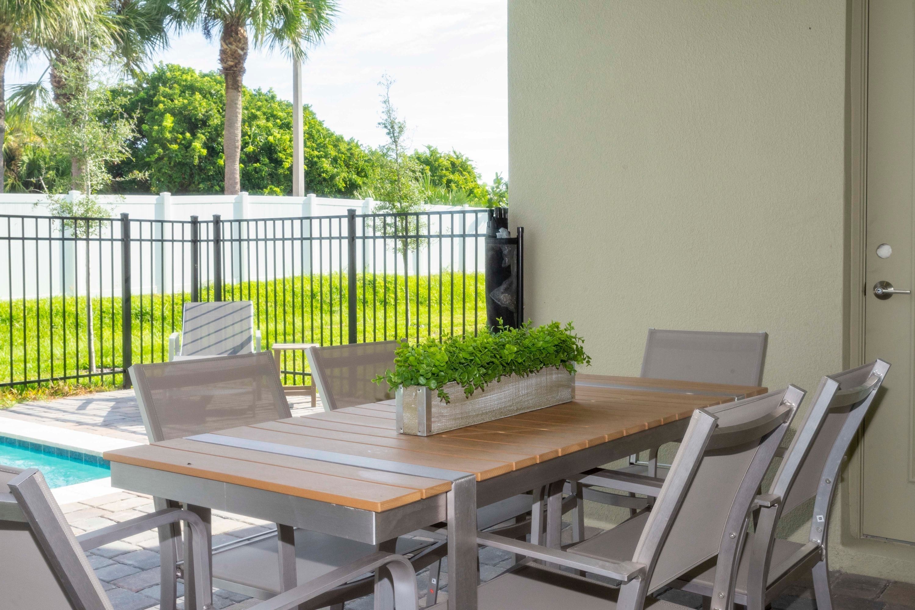 Een terras buiten met een houten tafel en stoelen, een plantenbak met groene planten, en een uitzicht op een zwembad en begroeiing.
