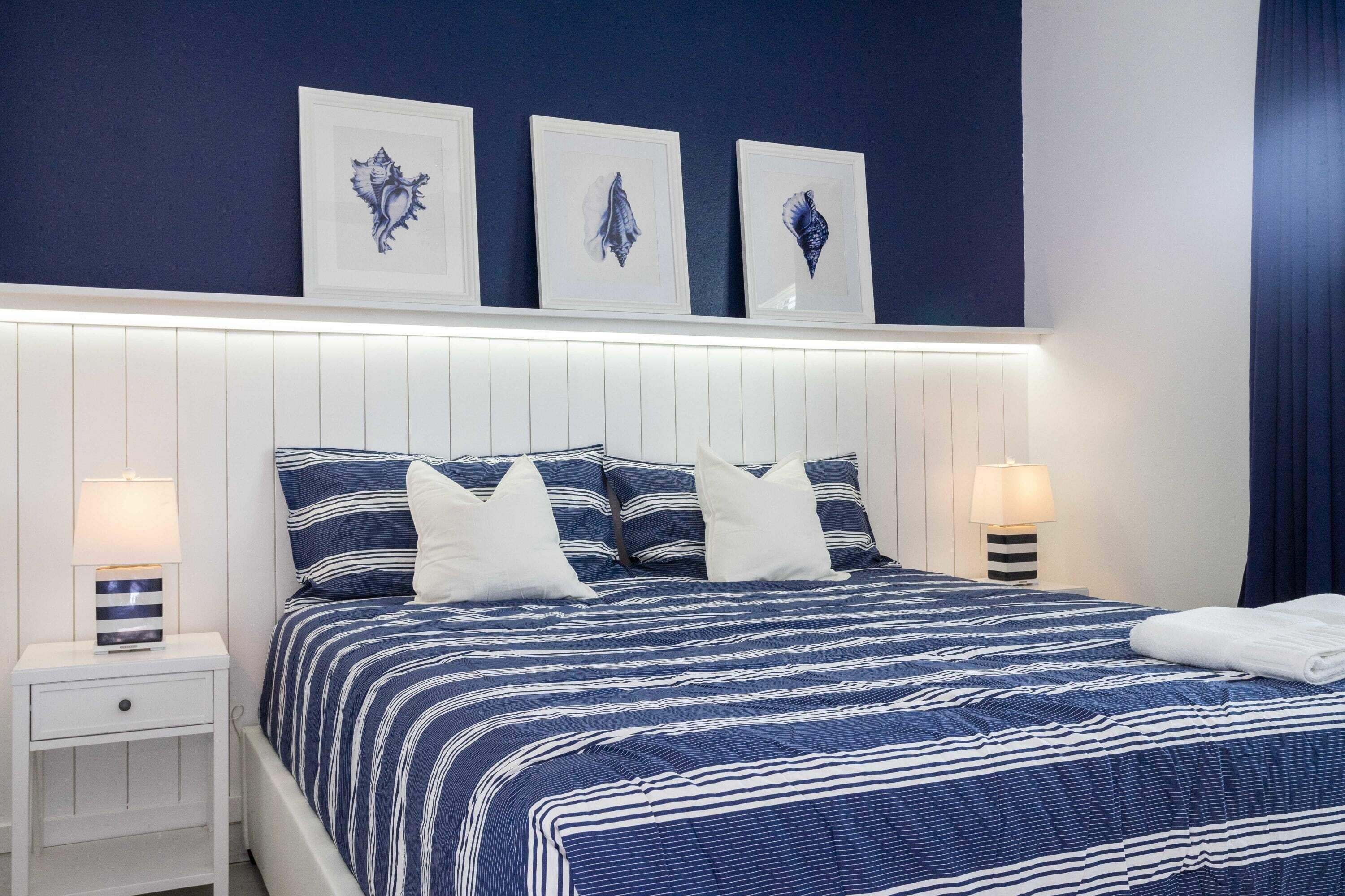 A neatly made bed with striped bedding, white pillows, and a blue headboard. There are framed pictures on the wall above the bed.